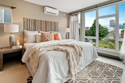 Photo of a bed with a white comforter, orange throw pillows, and a vertical striped headboard; one wall of all windows.